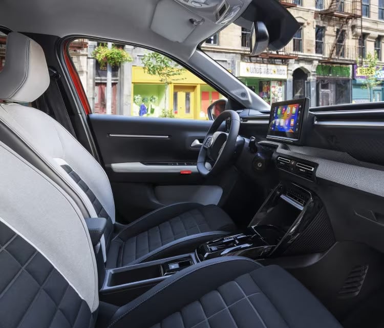 Auf diesem Bild ist der Innenraum eines modernen Autos mit seinen Vordersitzen, dem Armaturenbrett und einem großen Infotainmentsystem zu sehen, während man durch die Fenster eine städtische Umgebung erkennen kann.