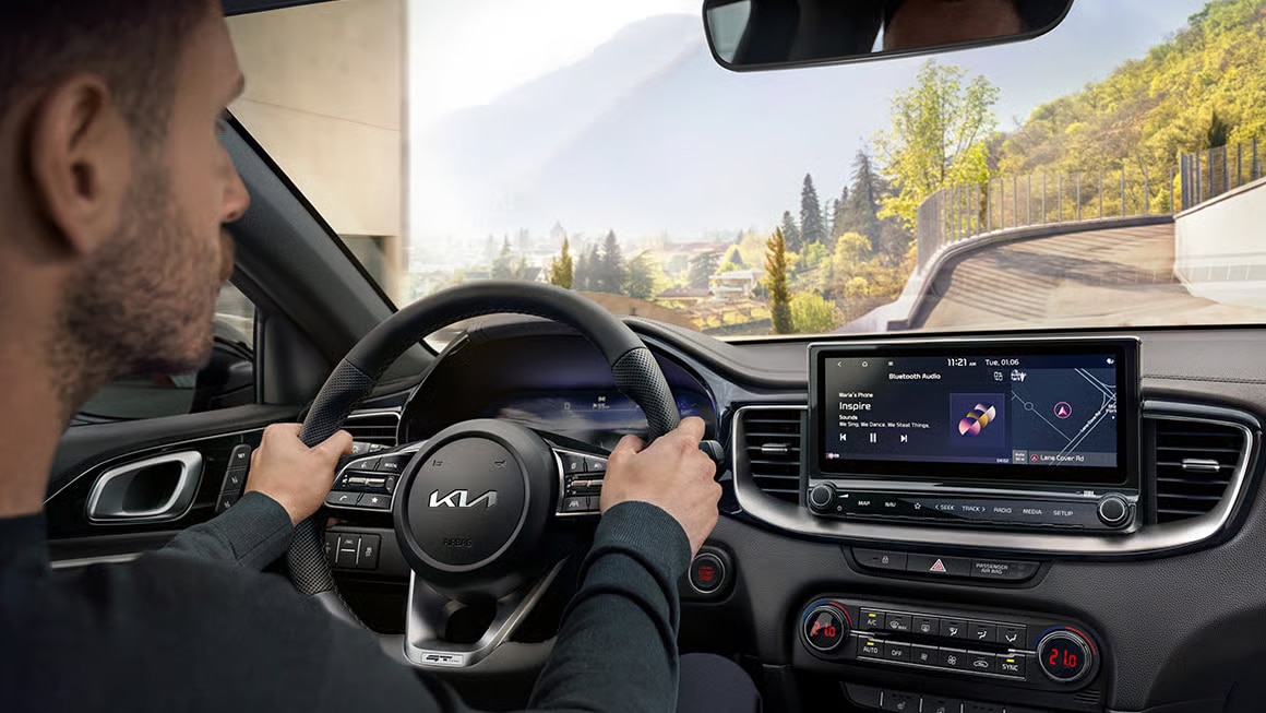 Das Bild zeigt einen Mann am Steuer eines Kia-Autos von innen, mit dem Blick auf das Infotainmentsystem, das Musik und Navigation anzeigt, und eine bergige Landschaft durch die Windschutzscheibe.