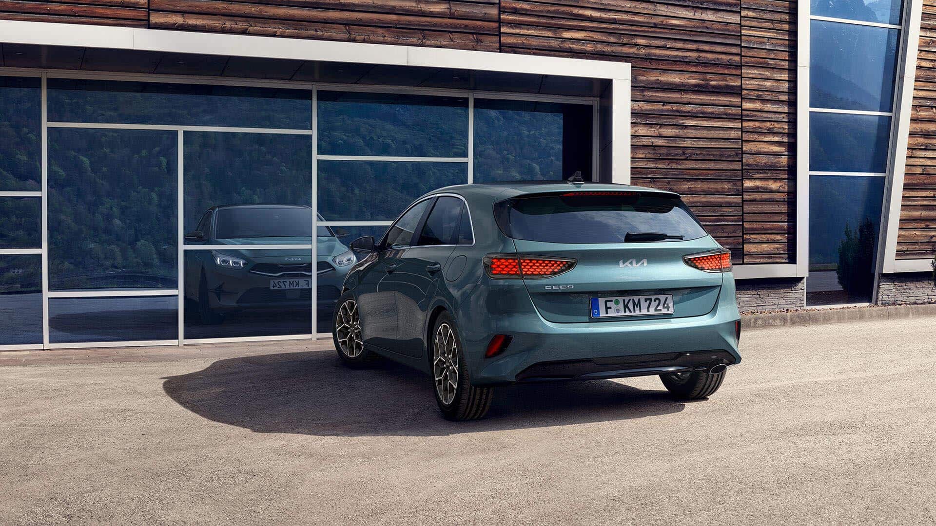 Auf diesem Bild ist ein türkisgrüner Kia Ceed mit deutschem Kennzeichen zu sehen, der vor einem modernen Gebäude mit Holzfassade und großen Glasfenstern parkt, in denen sich Bäume und ein weiteres Auto spiegeln.
