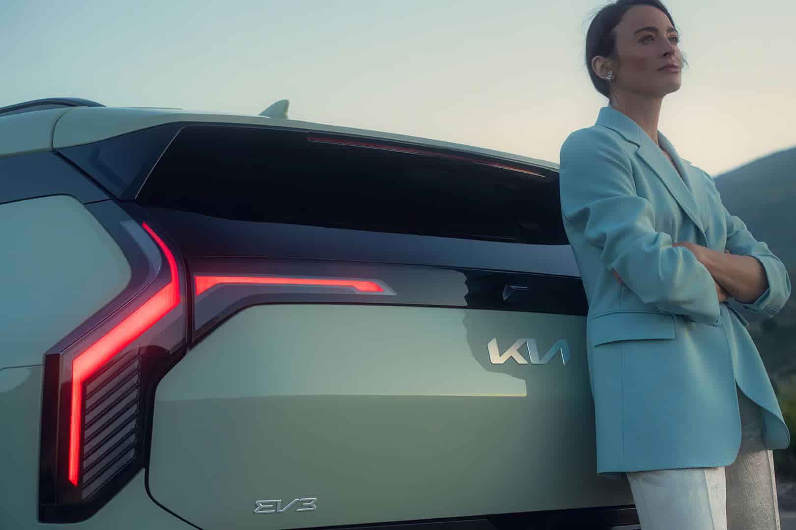 Auf diesem Bild ist eine Frau zu sehen, die im Freien an der hellgrünen Heckseite eines Kia EV3 lehnt, dessen Rücklichter leuchten.