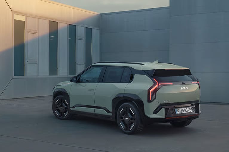 Auf diesem Bild ist ein moderner, hellgrüner Kia EV SUV in einer Dreiviertelansicht von hinten zu sehen, der vor einem minimalistischen Gebäude mit großen Fenstern geparkt ist.