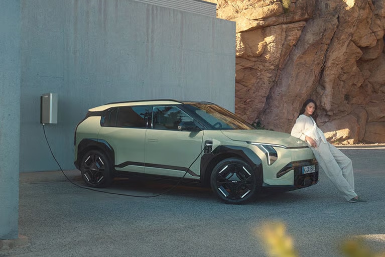 Ein hellgrünes Elektroauto wird an einer Wandladestation aufgeladen, während eine Frau lässig an der Front des Wagens lehnt, vor dem Hintergrund einer modernen Wand und natürlichen Felsen.