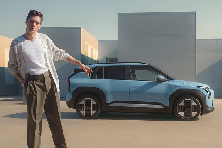 Ein Mann in einer hellen Strickjacke und dunklen Hosen zeigt auf einen hellblauen SUV, der vor modernen Gebäuden steht.