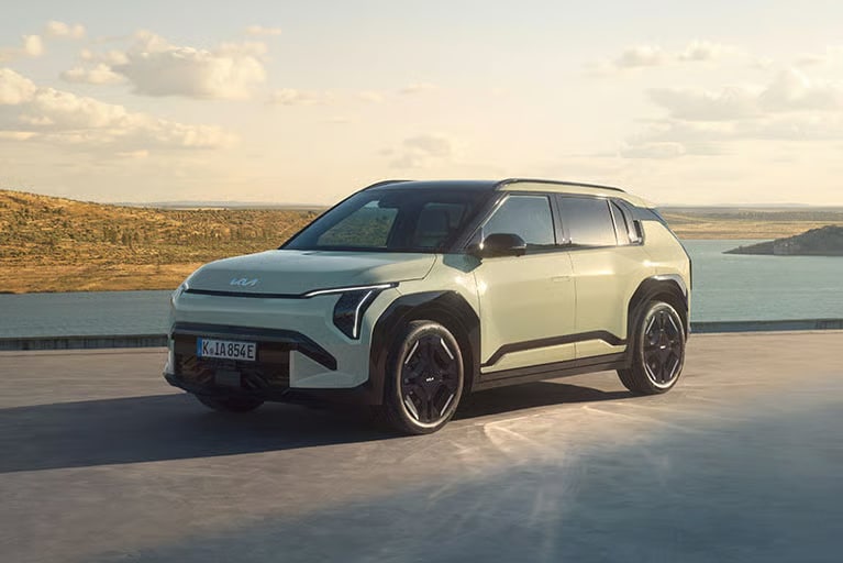 Auf diesem Bild ist ein beigefarbener Kia Elektro-SUV zu sehen, der vor einer malerischen Landschaft mit einem großen See und hügeligen Ufern parkt.