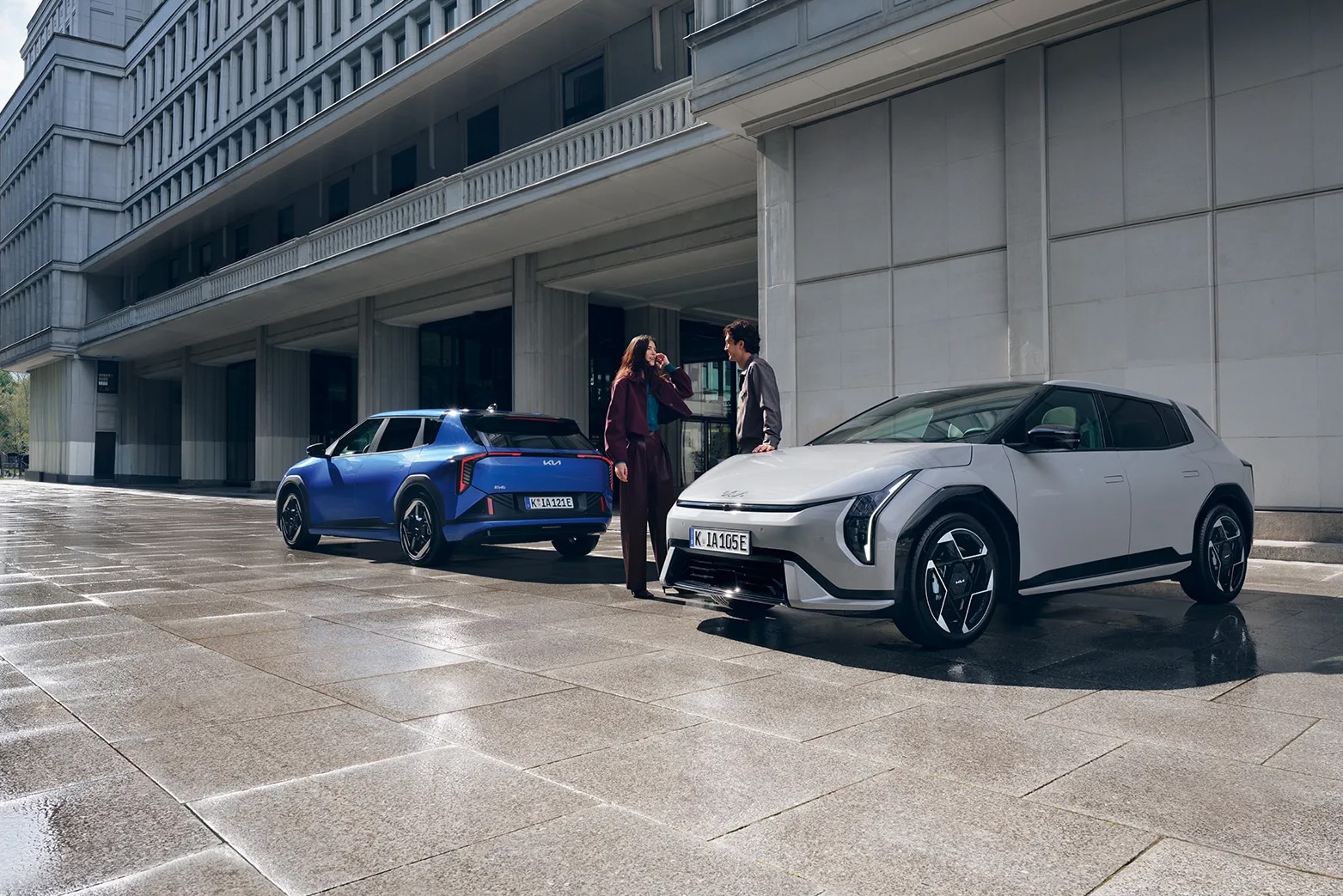 Das Bild zeigt zwei Kia-Fahrzeuge, ein blaues und ein hellgraues, mit einem Mann und einer Frau, die auf einem gepflasterten Platz vor einem modernen Gebäude stehen.