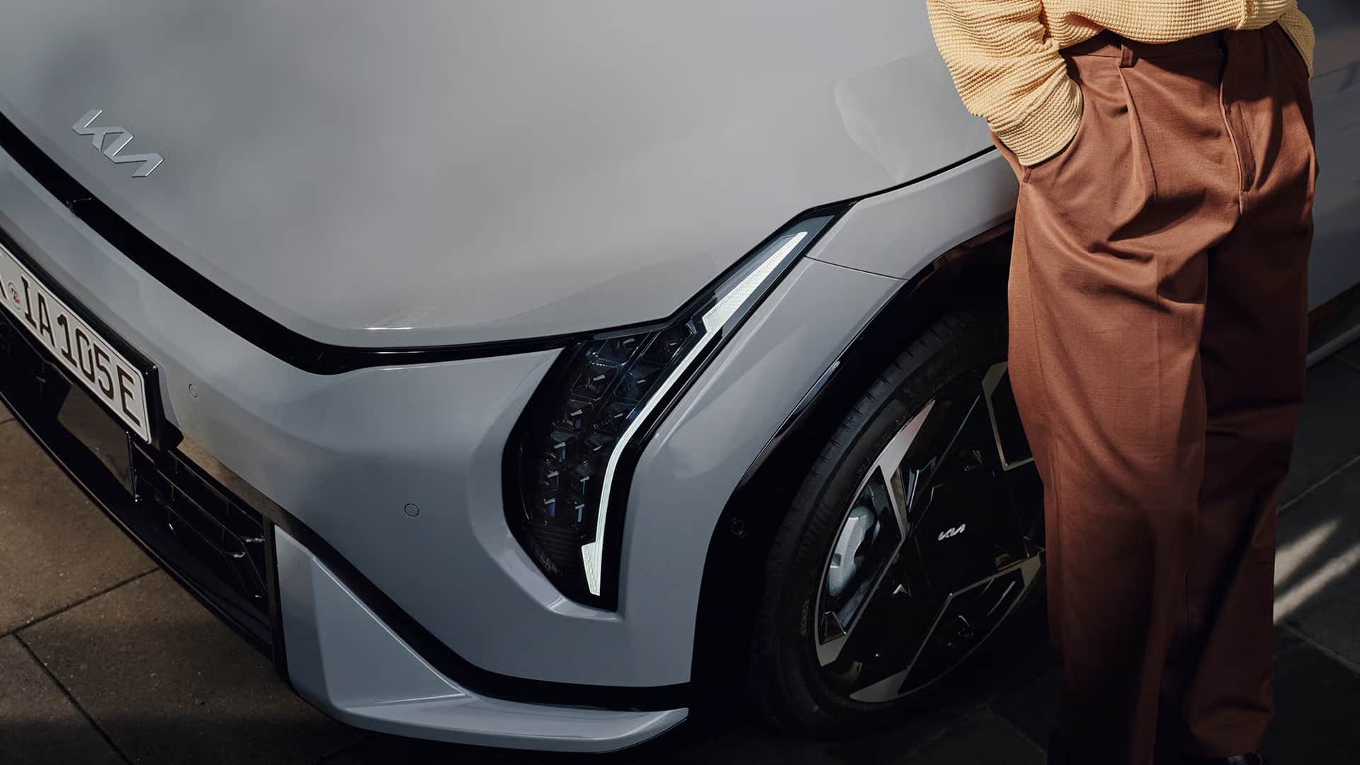 Auf diesem Bild ist die Front eines grauen Kia-Autos mit leuchtenden Scheinwerfern zu sehen, daneben steht eine Person in brauner Hose und hellem Pullover.