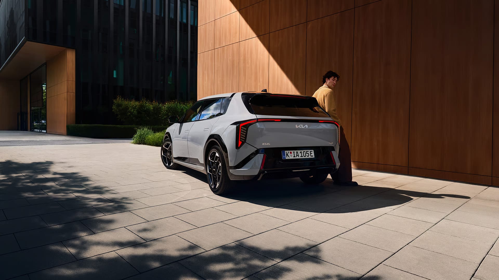 Auf diesem Bild ist ein grauer Kia EV6 mit einem Mann, der danebensteht, auf einem gepflasterten Platz vor modernen Gebäuden mit Holzfassade und dunklen Fenstern zu sehen.