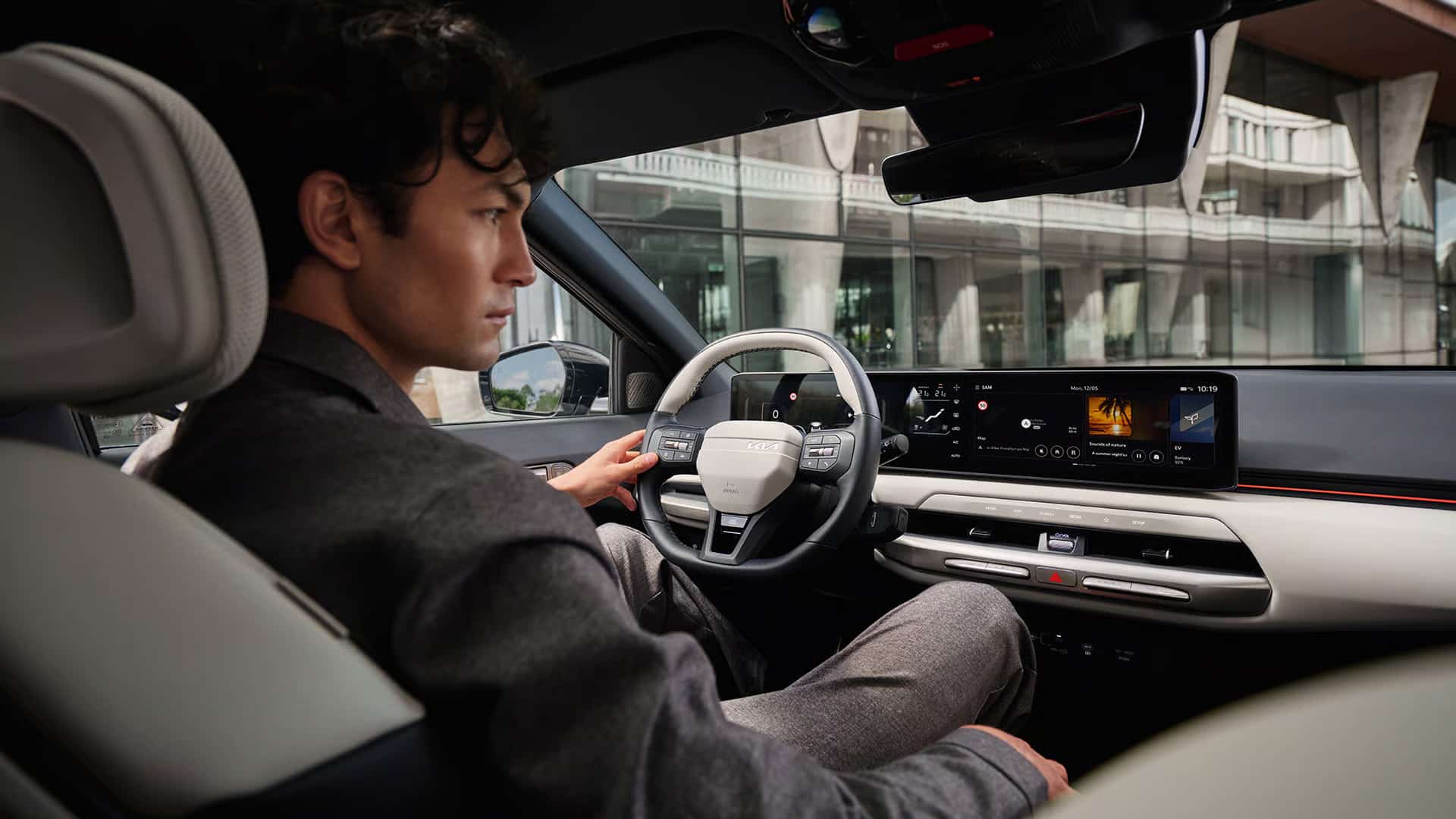 Ein Mann sitzt am Steuer eines modernen Autos mit einem digitalen Display und einem markanten Lenkrad, während im Hintergrund ein modernes Gebäude mit Glasfassade zu sehen ist.