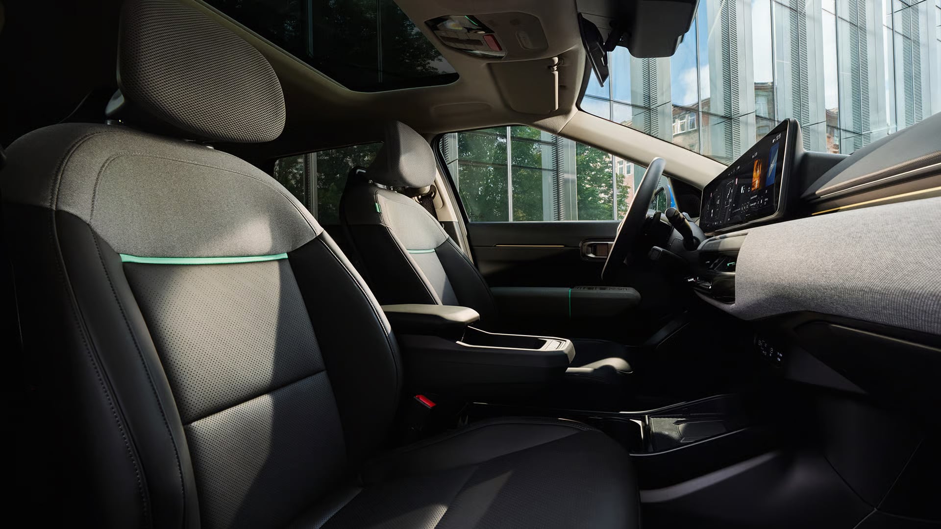 Das Bild zeigt den modernen Innenraum eines Autos, der Sitze aus Stoff und perforiertem Leder mit einem grünen Akzentstreifen, ein großes digitales Display auf dem Armaturenbrett sowie ein Panorama-Glasdach umfasst, mit Blick auf eine moderne Glasfassade und Bäume außerhalb des Fahrzeugs.