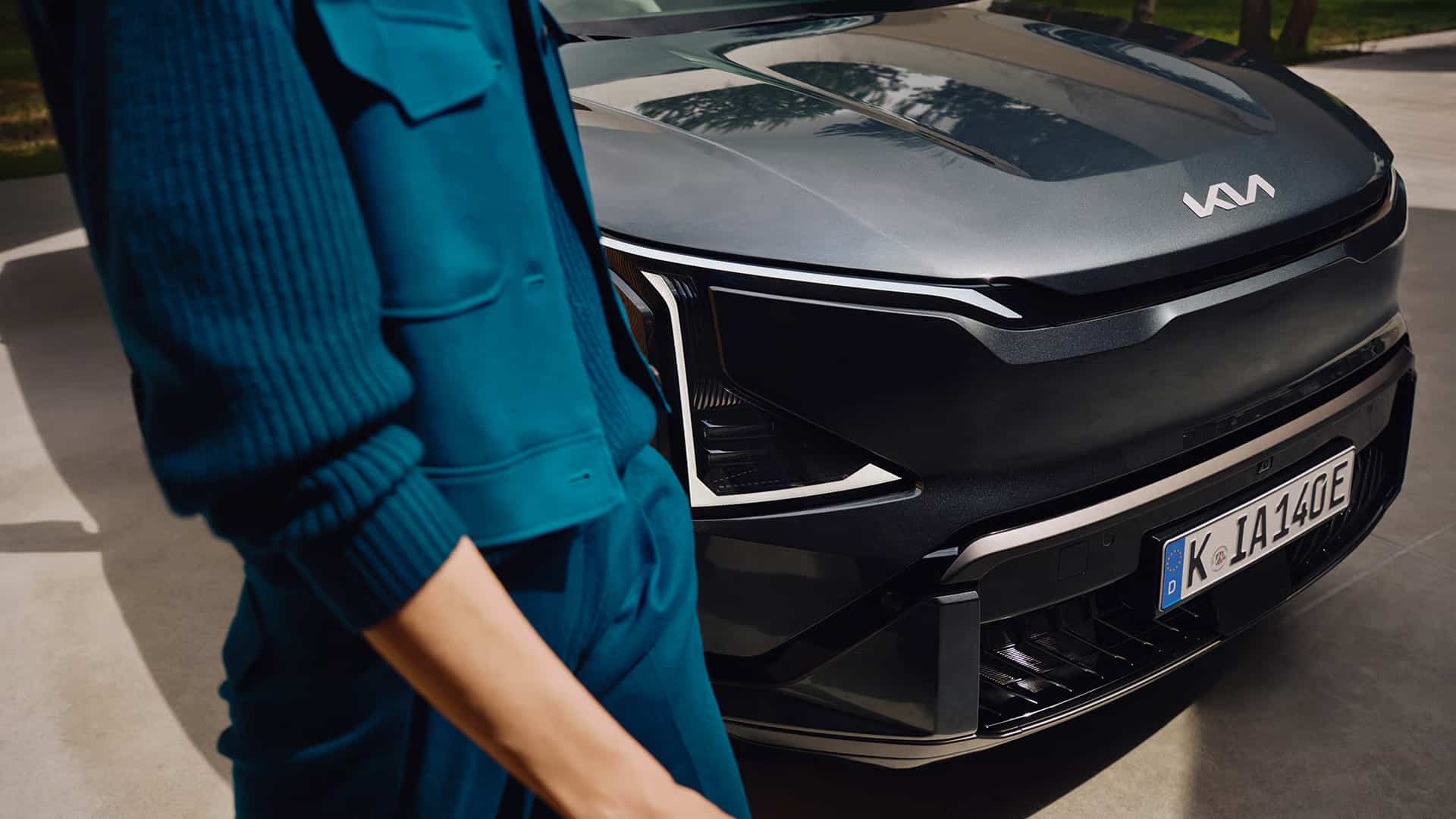 Auf diesem Bild sind die Front eines dunklen Kia-Fahrzeugs mit deutschem Nummernschild und eine Person in türkiser Kleidung zu sehen.