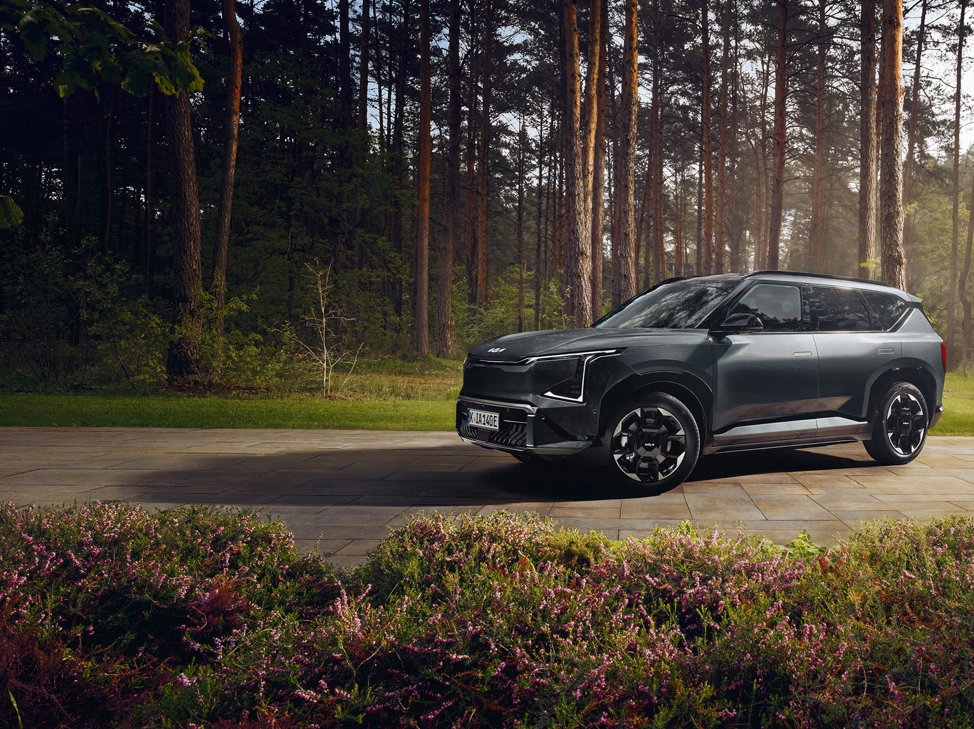 Auf diesem Bild ist ein modernes, dunkelgraues SUV zu sehen, das auf einem gepflasterten Weg vor einem Kiefernwald parkt und im Vordergrund von blühenden Sträuchern umgeben ist.