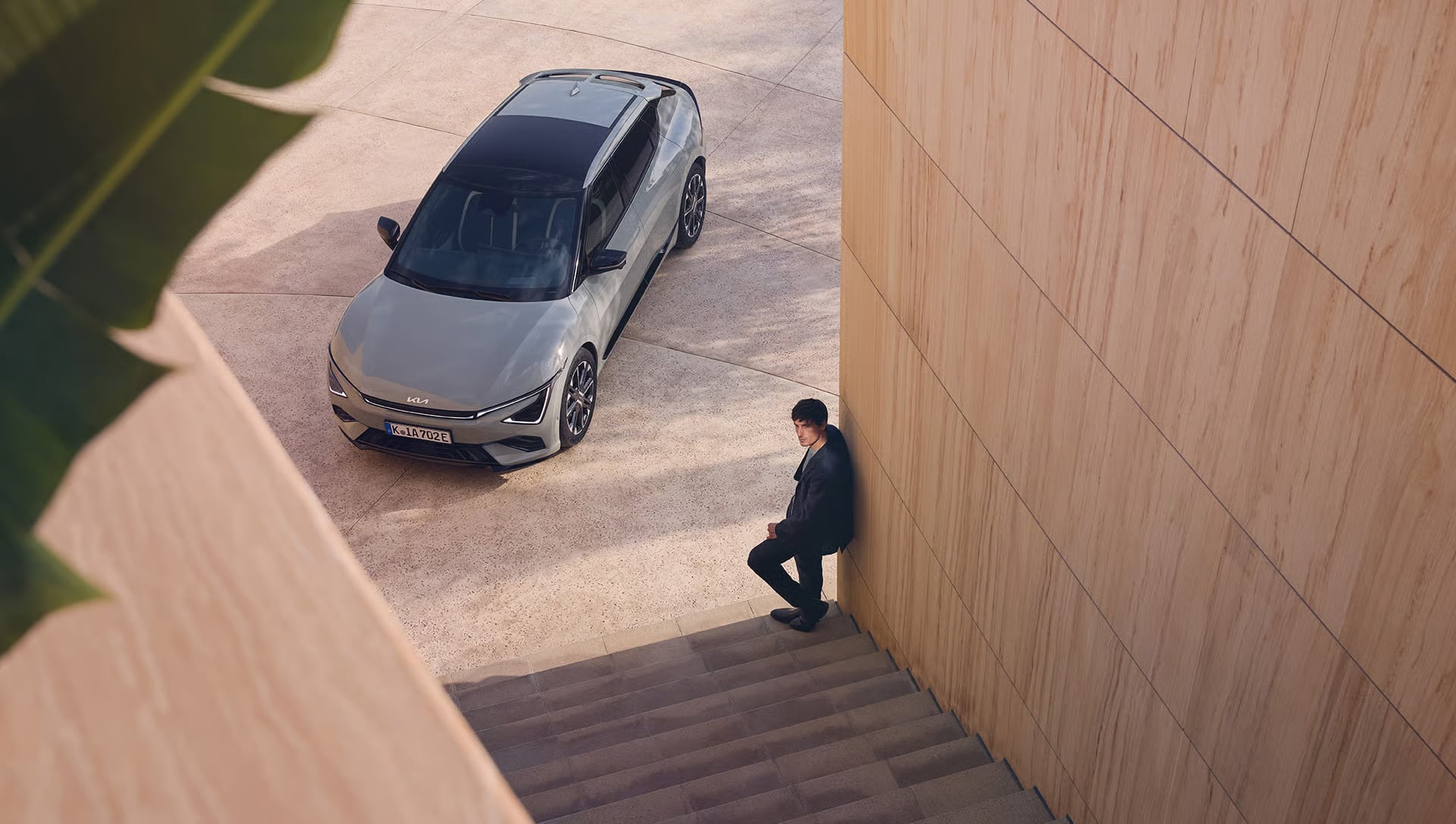 Das Bild zeigt ein modernes graues Auto auf einem gepflasterten Vorplatz und einen Mann, der an einer holzverkleideten Wand lehnt und auf Stufen sitzt.