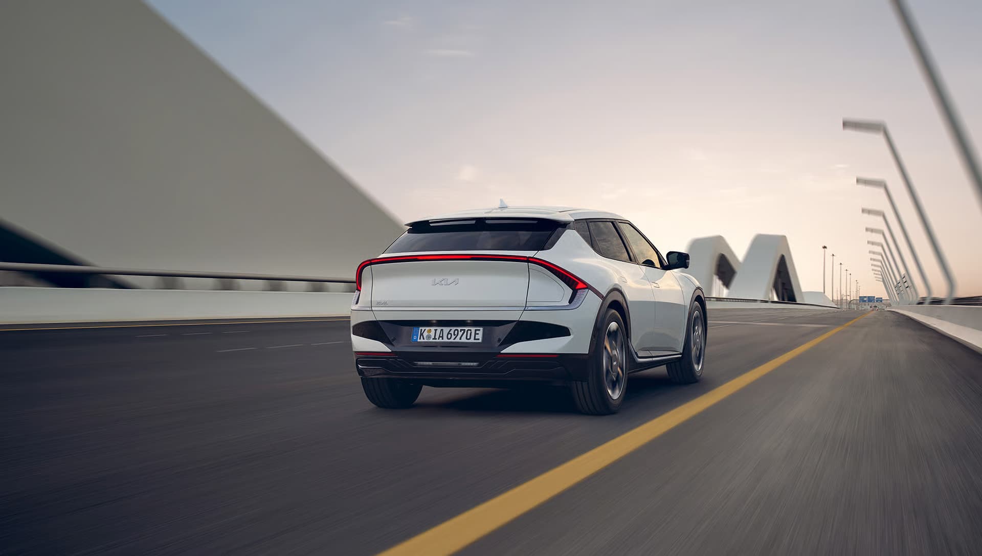 Auf diesem Bild ist ein weißer Kia EV6 von hinten zu sehen, der bei Dämmerung auf einer modernen Brückenstraße mit markanter Architektur im Hintergrund fährt.