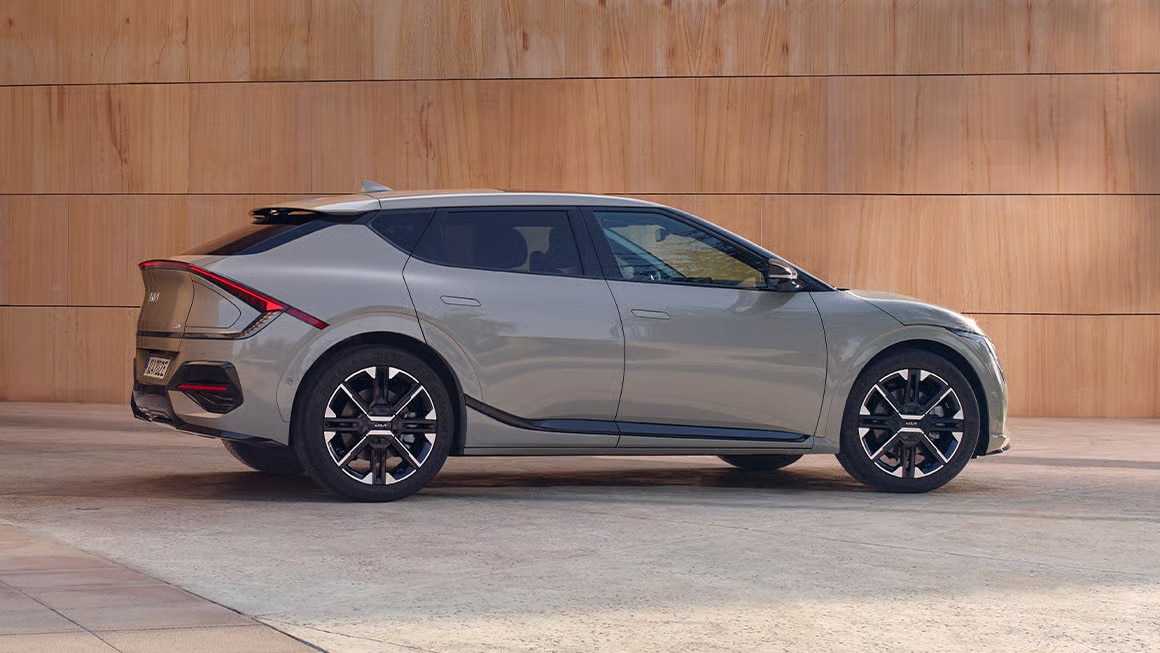 Auf diesem Bild ist ein hellgrauer Kia EV6 von der Seite vor einer Holzwand geparkt zu sehen.