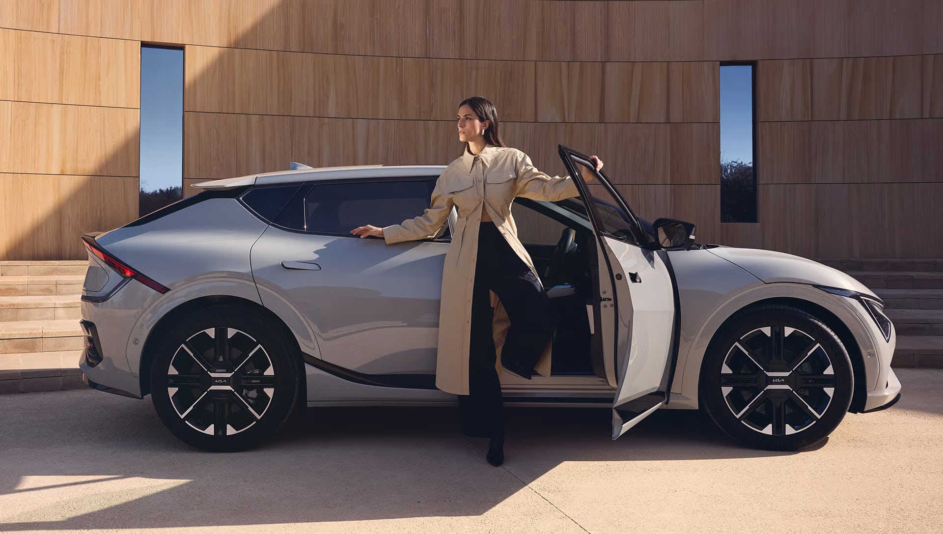 Auf diesem Bild ist eine Frau in einem beigefarbenen Mantel zu sehen, die neben einem modernen, hellgrauen Auto mit offener Tür vor einem Gebäude mit Holzfassade posiert.