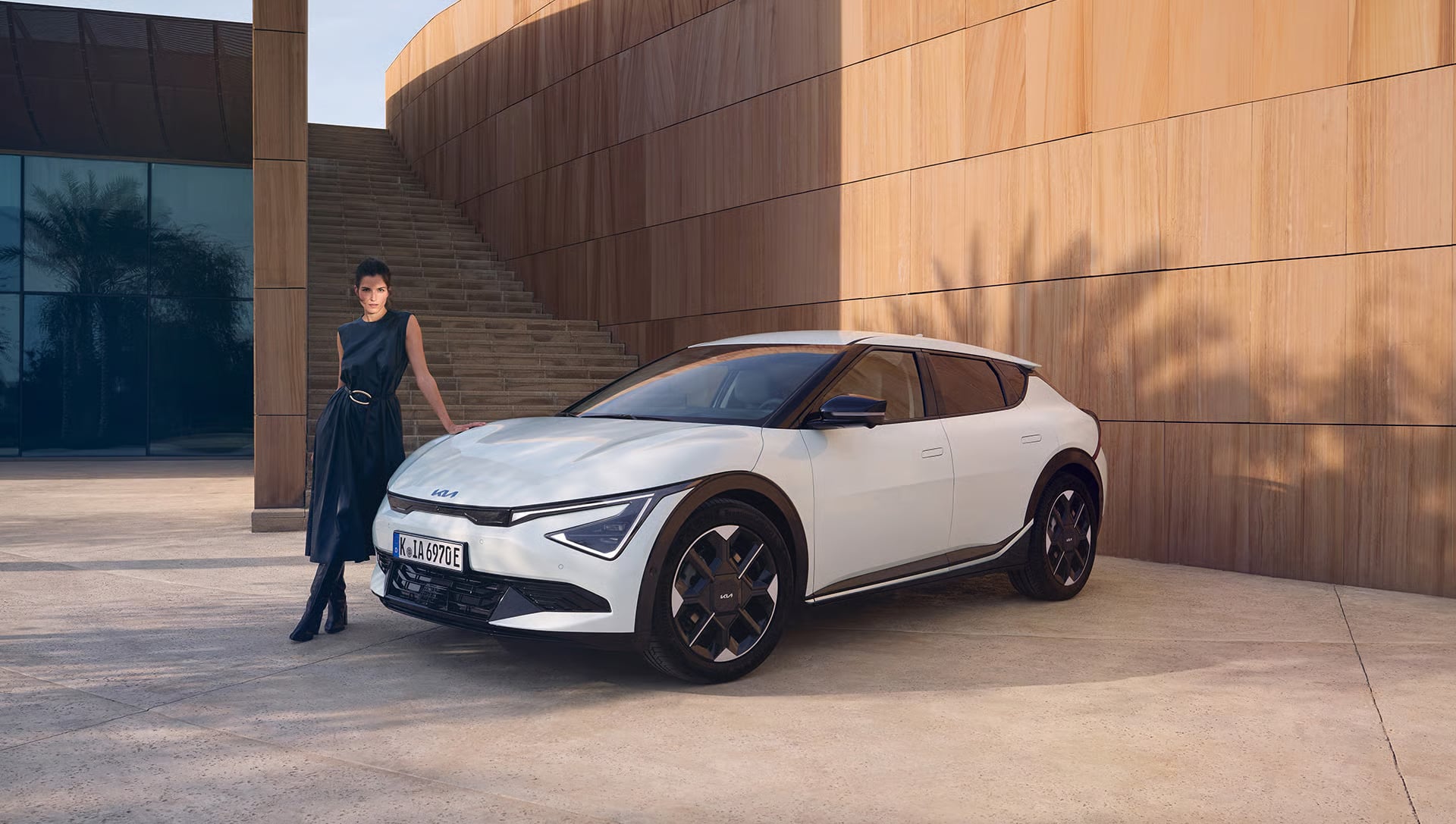 Auf diesem Bild ist eine elegant gekleidete Frau zu sehen, die an einem weißen Kia EV6 vor einer modernen Architektur mit Holzfassaden und einer Glasfront lehnt.