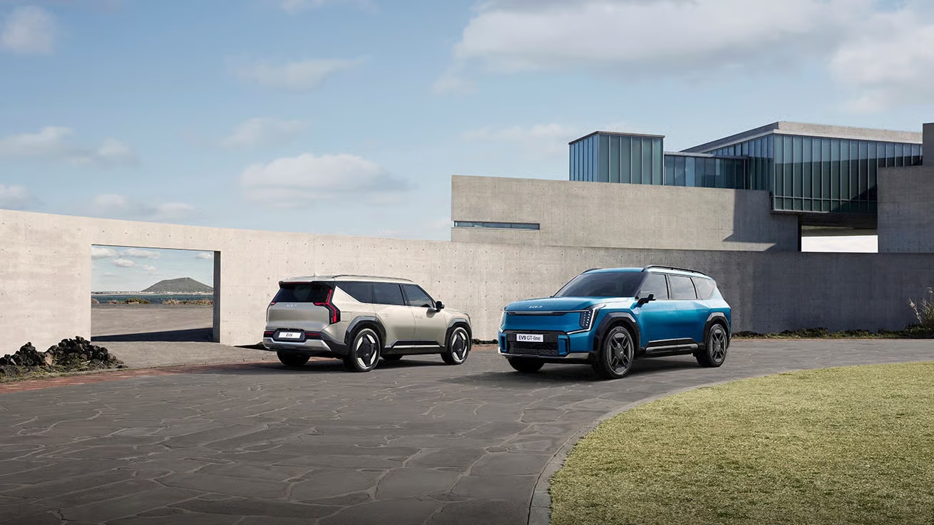 Auf diesem Bild sind zwei Kia EV9 SUVs vor einem modernen Gebäude mit Blick auf eine Küstenlandschaft durch eine Wandöffnung zu sehen.