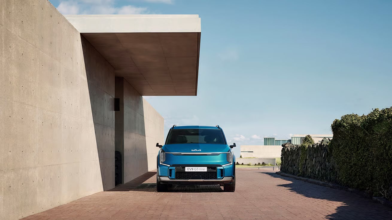 Auf diesem Bild ist ein blaues Kia EV9 GT-line Fahrzeug vor einer modernen Betonarchitektur auf einem gepflasterten Weg unter blauem Himmel zu sehen.