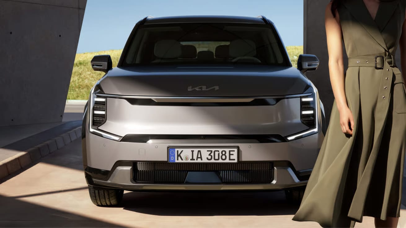 Auf diesem Bild sind die Front eines modernen, grauen Kia SUV mit deutschem Kennzeichen und eine danebenstehende Frau in einem olivgrünen Kleid vor einem Gebäude in heller Außenumgebung zu sehen.