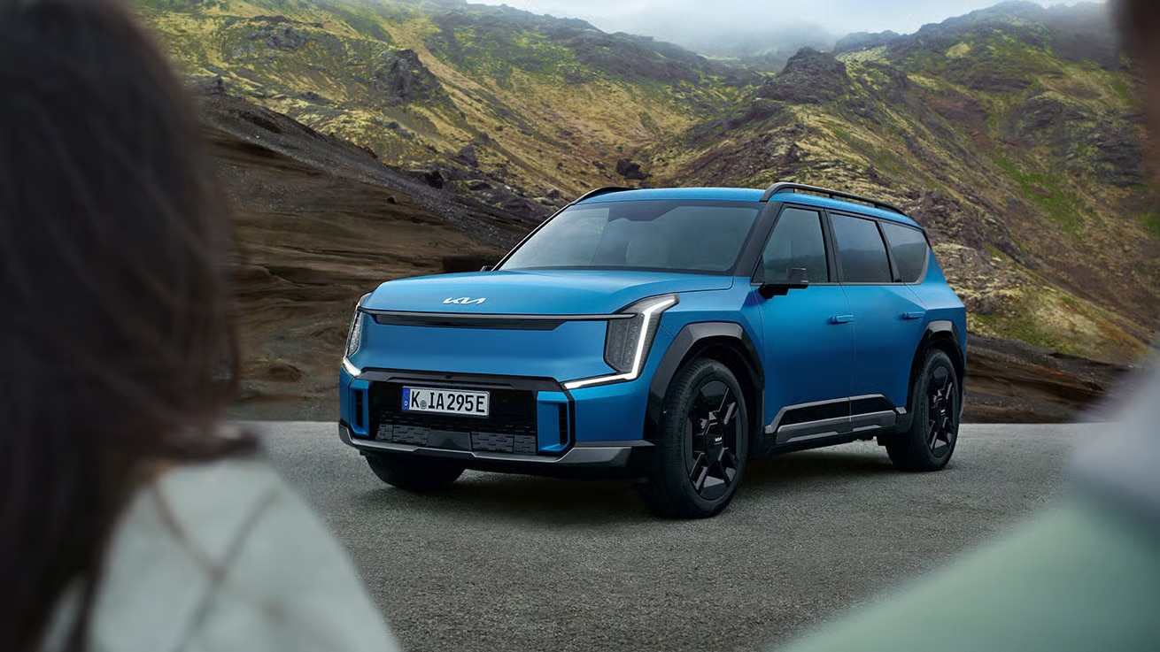 Auf diesem Bild ist ein blauer Kia EV9 SUV auf einer Straße in einer bergigen und kargen Landschaft zu sehen, eingerahmt von unscharfen Personen im Vordergrund.