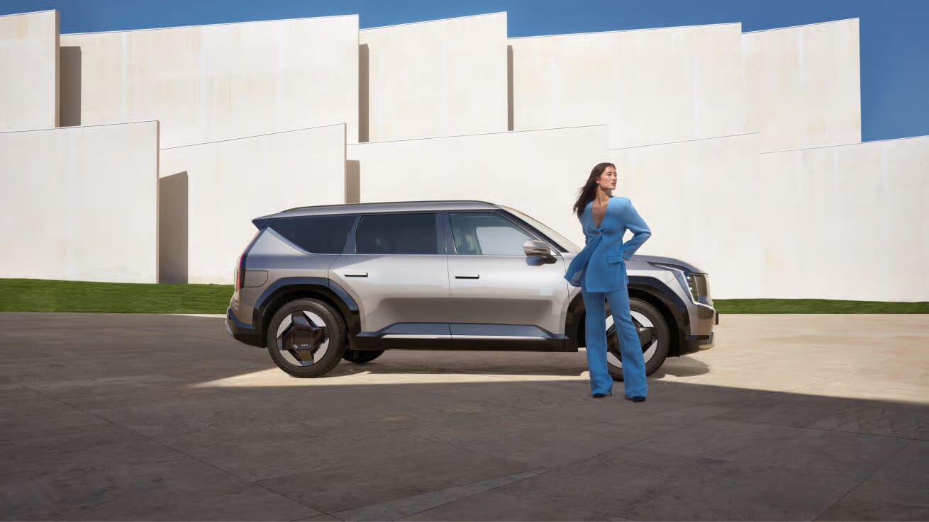 Das Bild zeigt eine Frau in einem blauen Anzug, die neben einem modernen, grauen Auto vor einer hellen, geometrischen Gebäudekulisse unter blauem Himmel steht.