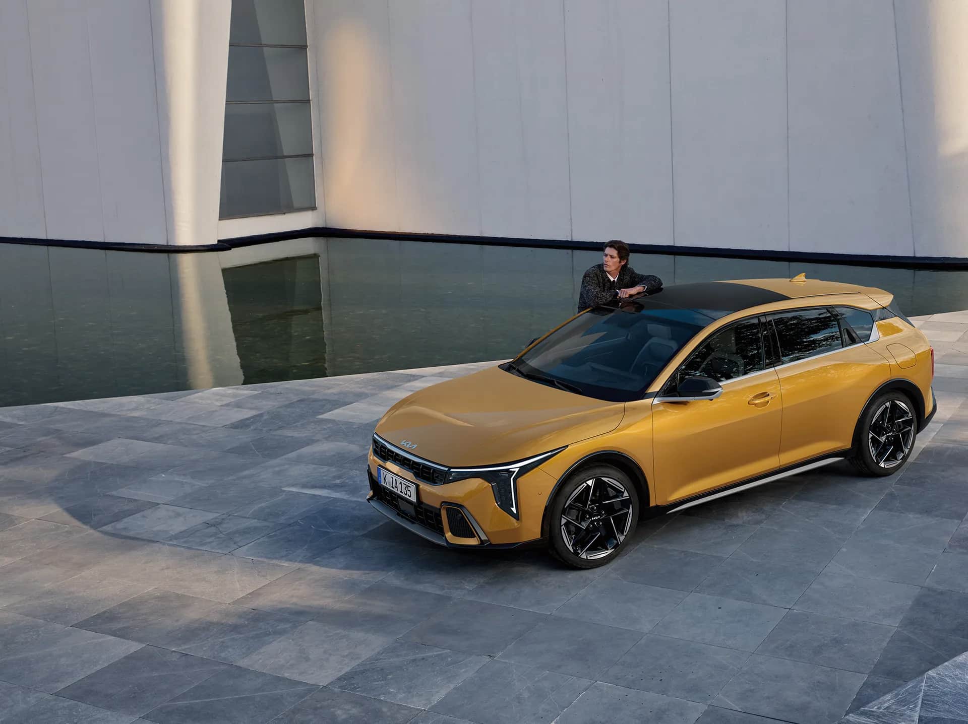 Auf diesem Bild ist ein goldgelbes Auto zu sehen, auf dessen Dach sich ein Mann lehnt, vor einer modernen weißen Gebäudefassade und einem Wasserbecken.