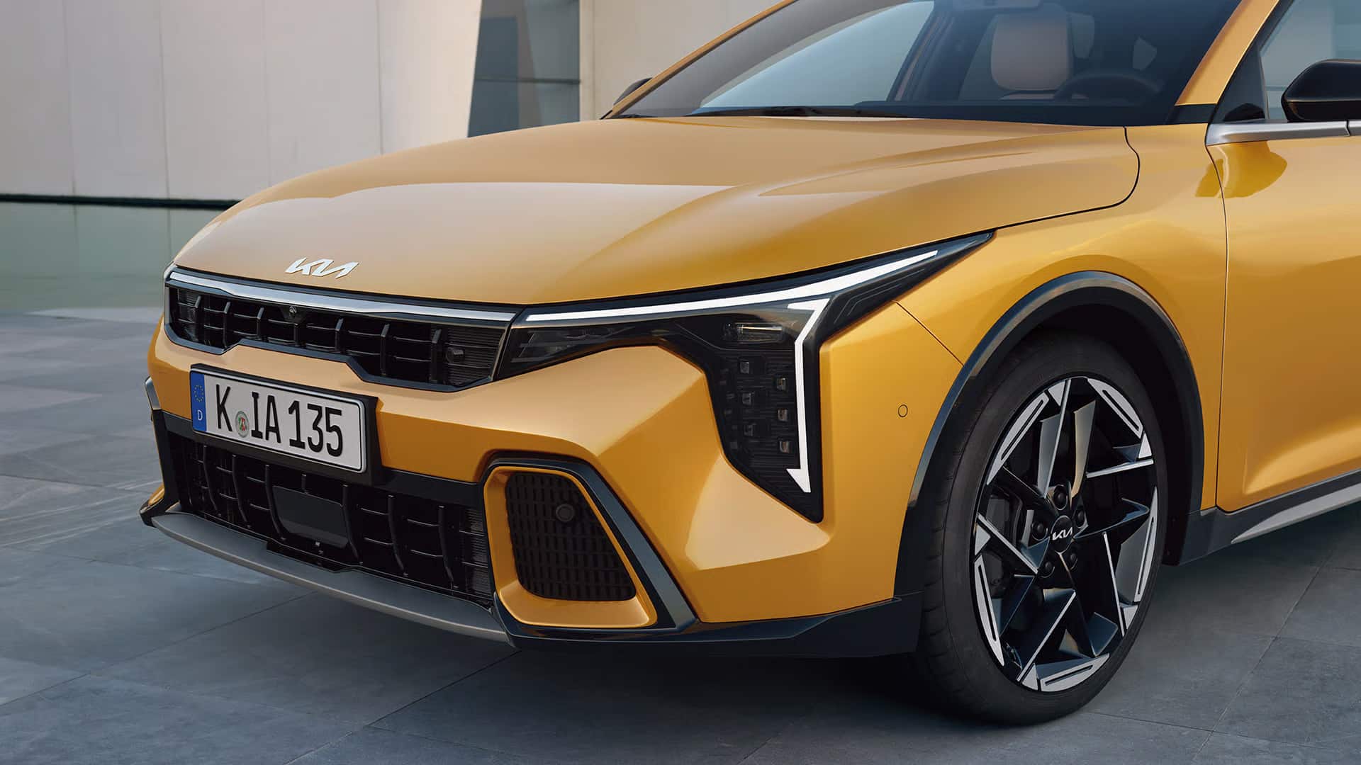 Auf diesem Bild ist die Frontpartie eines goldgelben Kia-Fahrzeugs mit auffälligen Scheinwerfern, einem deutschen Kennzeichen und einem Teil des Vorderrades zu sehen.