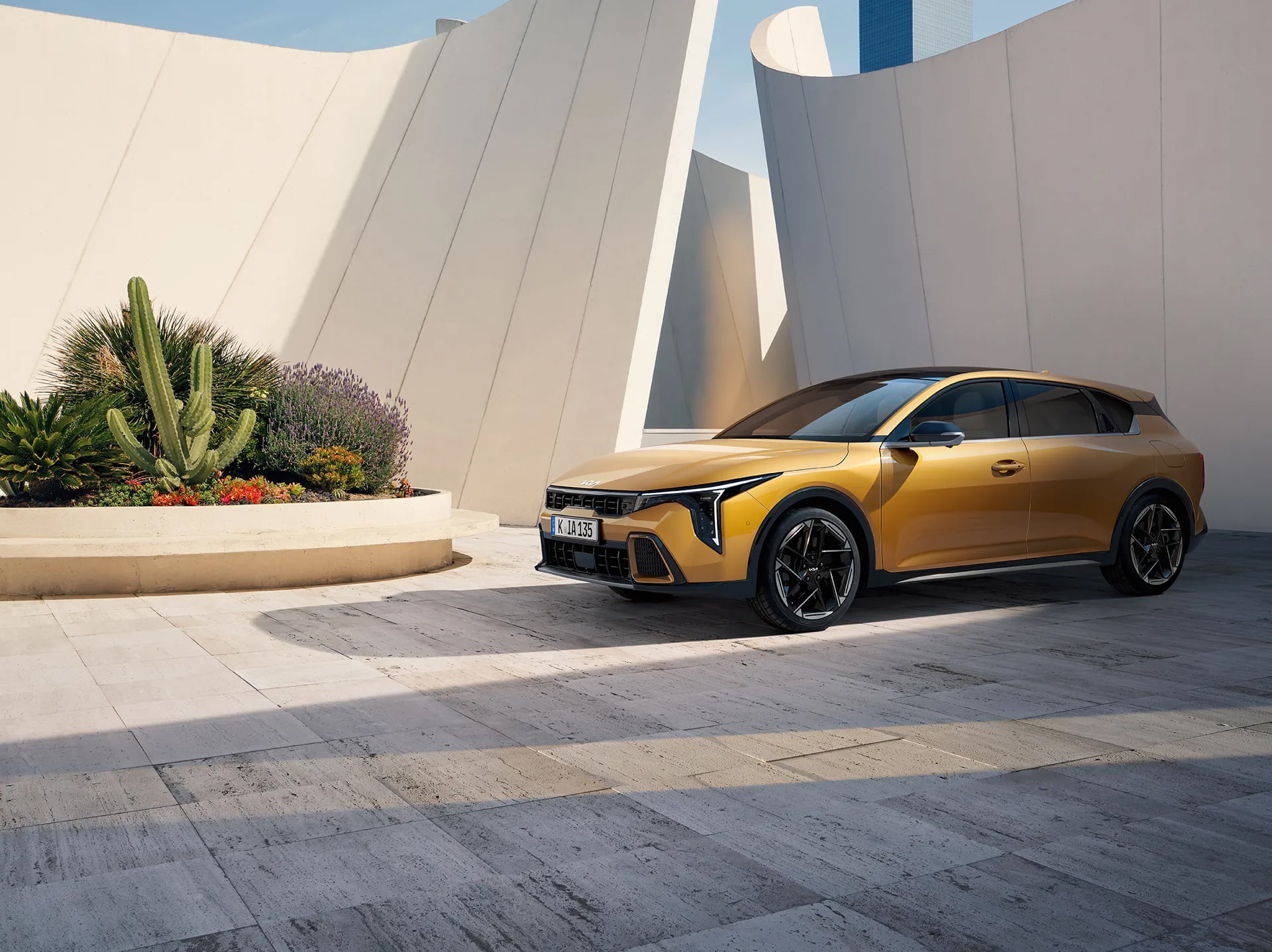 Auf dem Bild ist ein modernes, goldfarbenes Auto zu sehen, das auf einem hellen Plattenweg vor einer markanten, geschwungenen Architektur mit Kakteen und blühenden Pflanzen steht, unter einem klaren blauen Himmel.