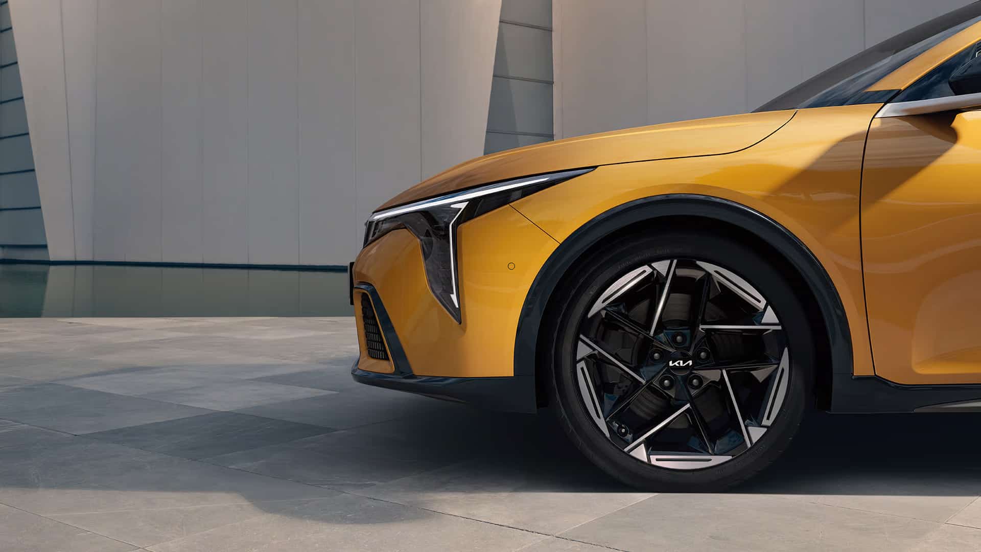 Auf diesem Bild ist die goldfarbene Front eines modernen Kia-Fahrzeugs mit einem Design-Rad vor einem hellen, architektonischen Gebäude zu sehen.