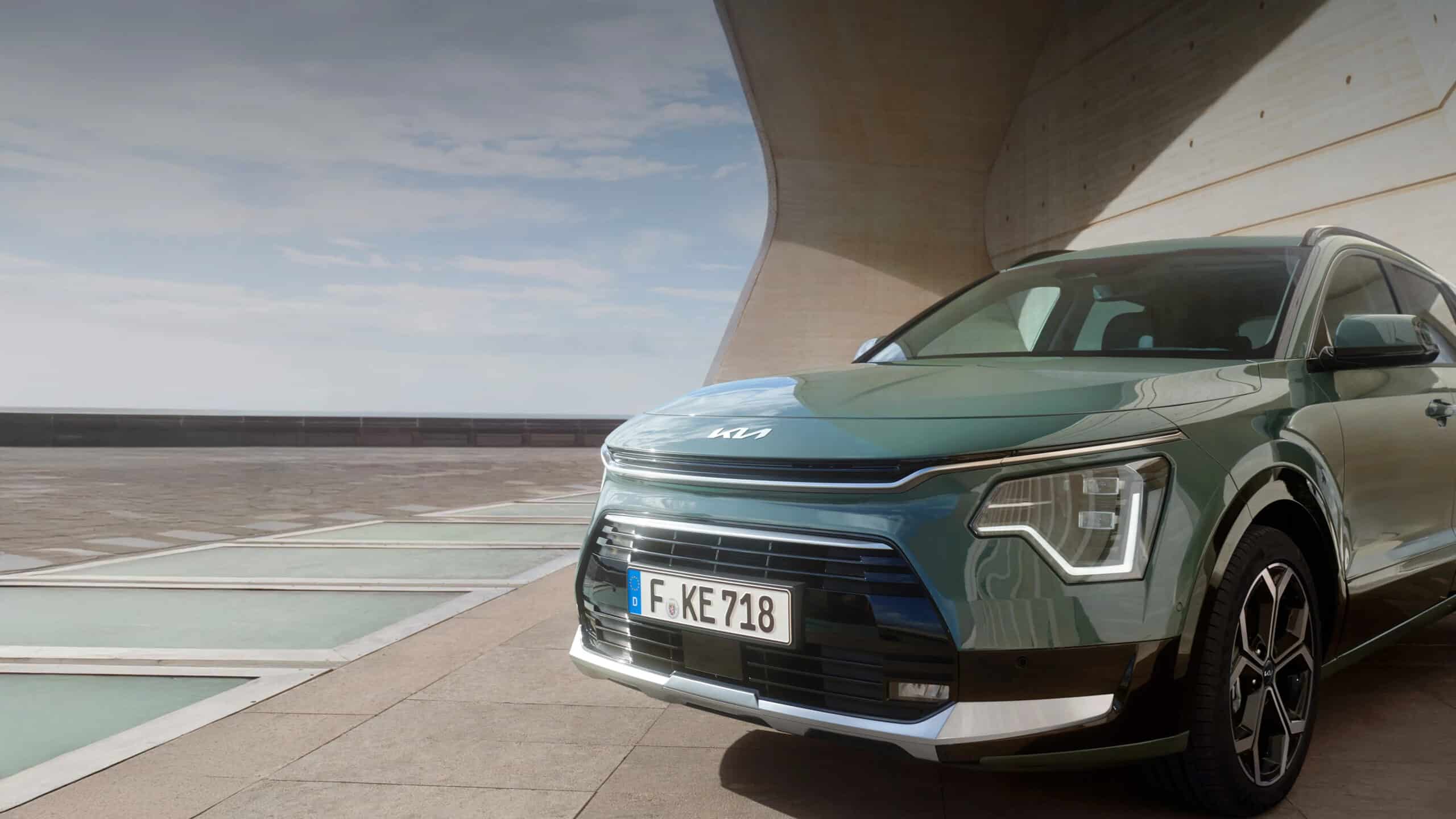 Auf diesem Bild ist ein grüner Kia Niro in einer modernen architektonischen Umgebung unter einem teilweise bewölkten Himmel mit Wasserflächen im Hintergrund zu sehen.