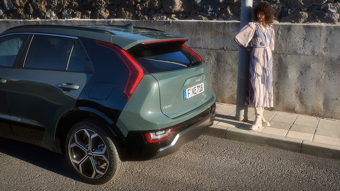 Auf diesem Bild ist ein grüner Kia Niro am Straßenrand zu sehen, daneben steht eine Frau auf dem Gehweg, die sich an einen Pfosten lehnt.