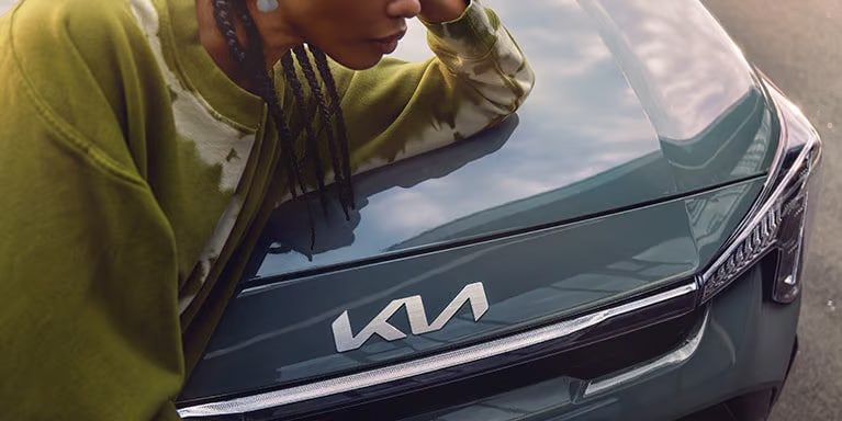 Auf dem Bild ist eine Person mit geflochtenen Haaren und einem grünen Batik-Sweatshirt zu sehen, die sich auf die Motorhaube eines dunkelgrünen Kia-Autos mit dem modernen Logo stützt.