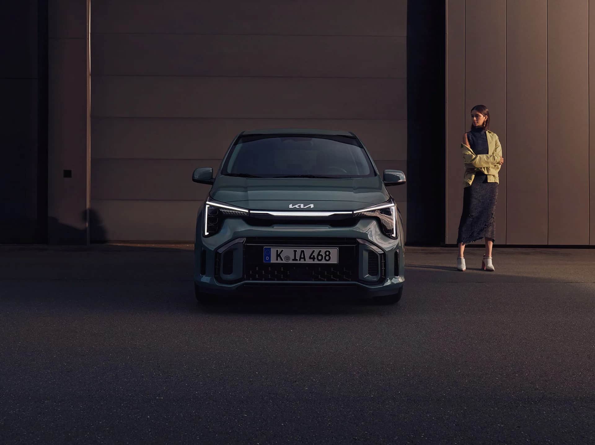 Auf diesem Bild sind ein grüner Kia-Kompaktwagen und eine stehende Frau vor einer dunklen, modernen Gebäudewand zu sehen.