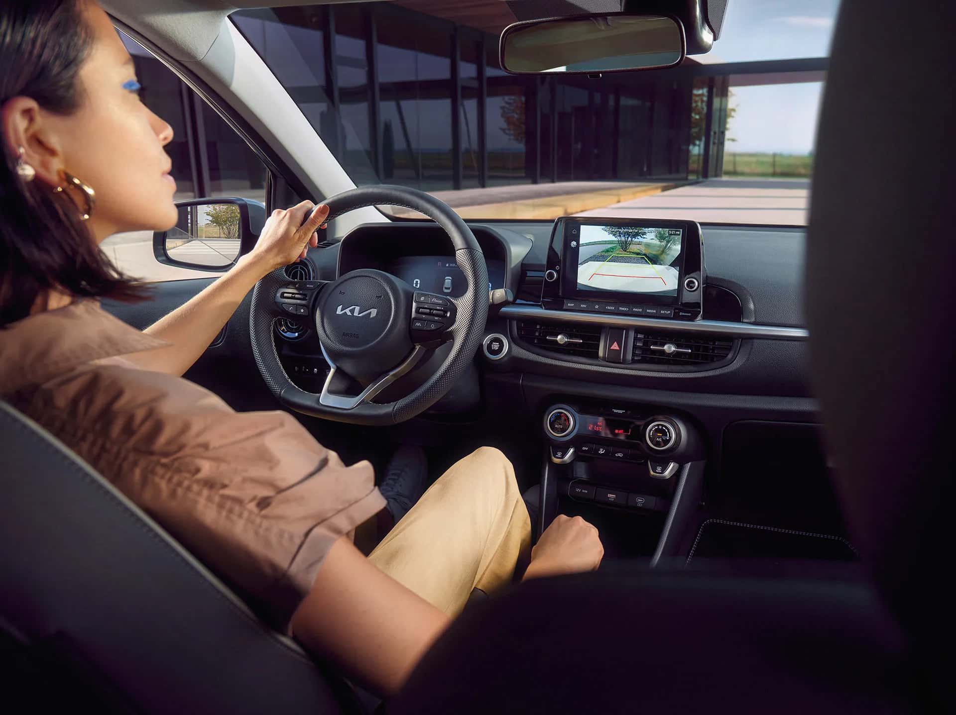 Auf diesem Bild ist eine Frau am Steuer eines modernen Kia-Autos zu sehen, dessen Innenraum und eine aktive Rückfahrkamera auf dem Display erkennbar sind.