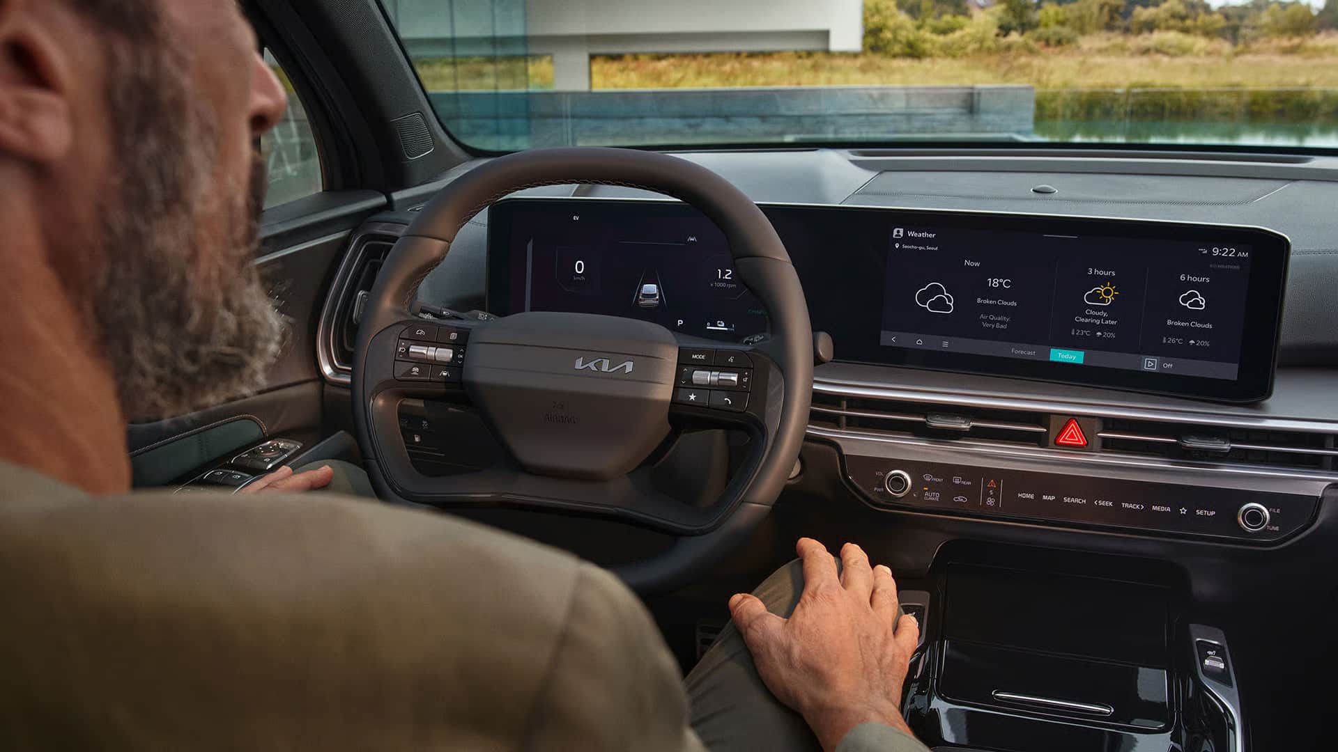 Das Bild zeigt den Innenraum eines modernen Autos mit einem Fahrer im Vordergrund, der das Lenkrad und die digitalen Displays sowie die Mittelkonsole betrachtet, während im Hintergrund eine Landschaft mit einem Gebäude zu sehen ist.