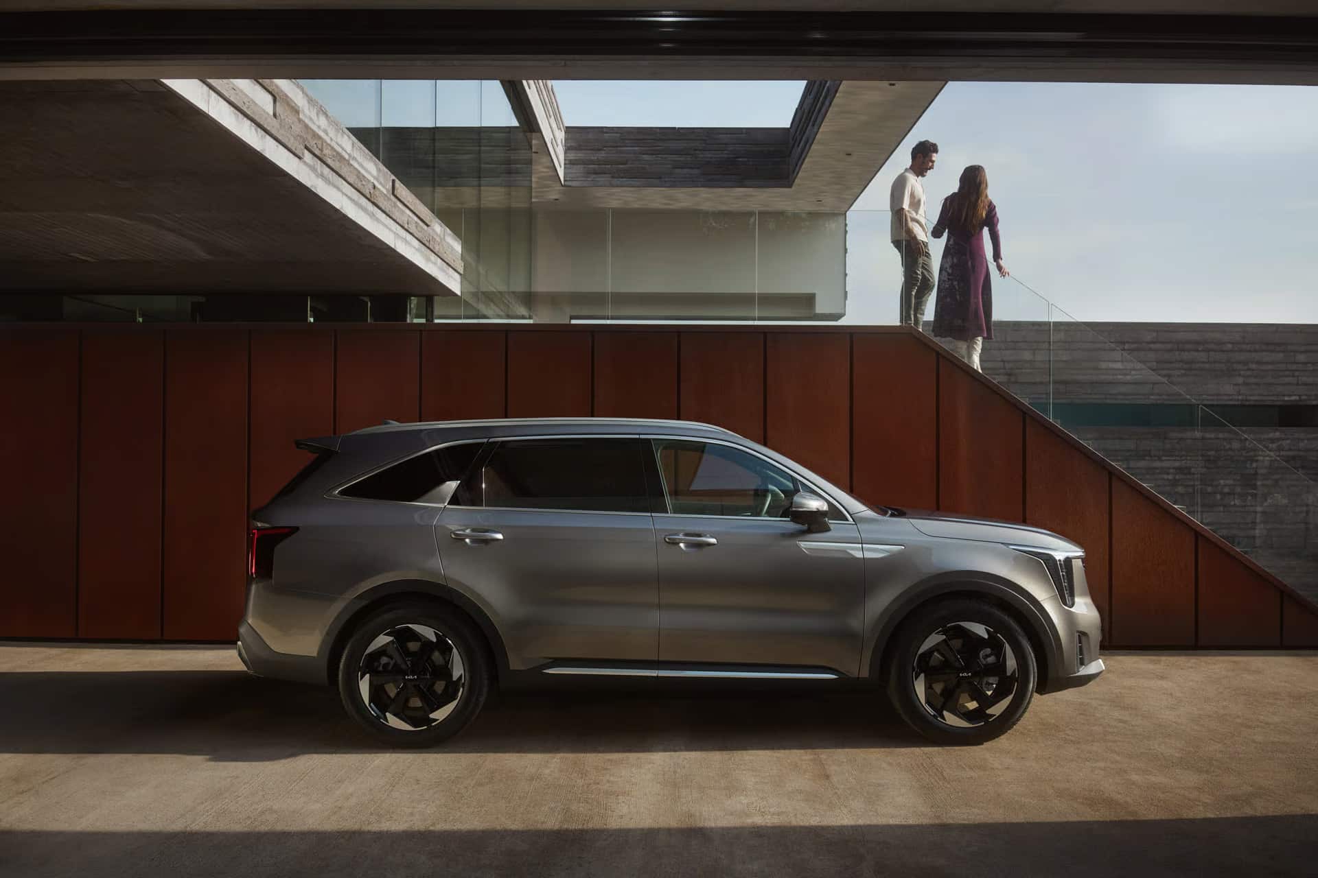 Das Bild zeigt einen grauen SUV, der vor einem modernen Gebäude mit rostroten Wänden und Glasflächen parkt, während darüber ein Mann und eine Frau auf einer erhöhten Ebene stehen.