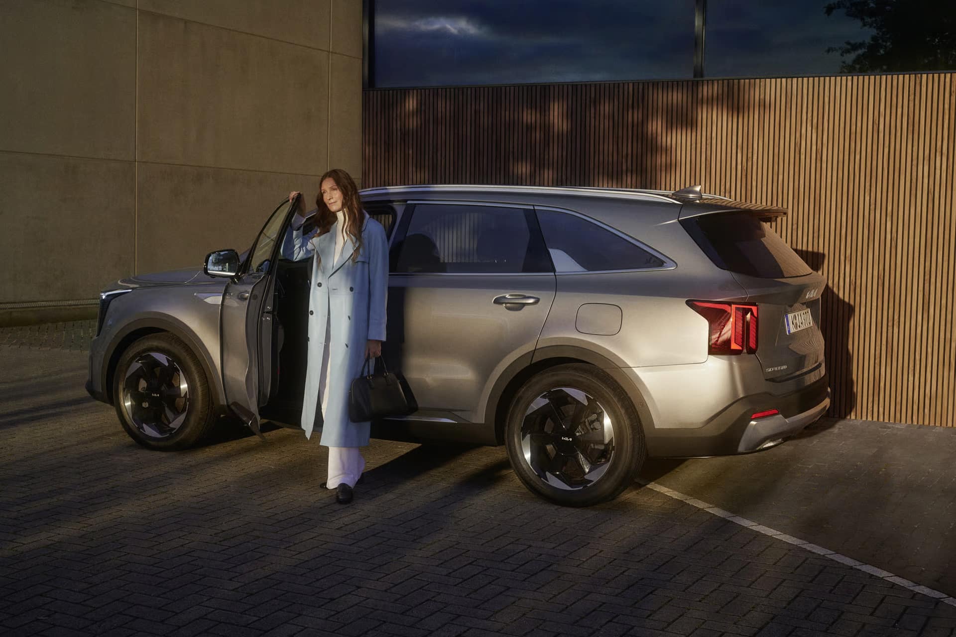 Auf diesem Bild ist eine Frau mit einer Handtasche zu sehen, die neben einem grauen Kia Sorento mit geöffneter Fahrertür auf einem gepflasterten Parkplatz vor einem modernen Gebäude steht.