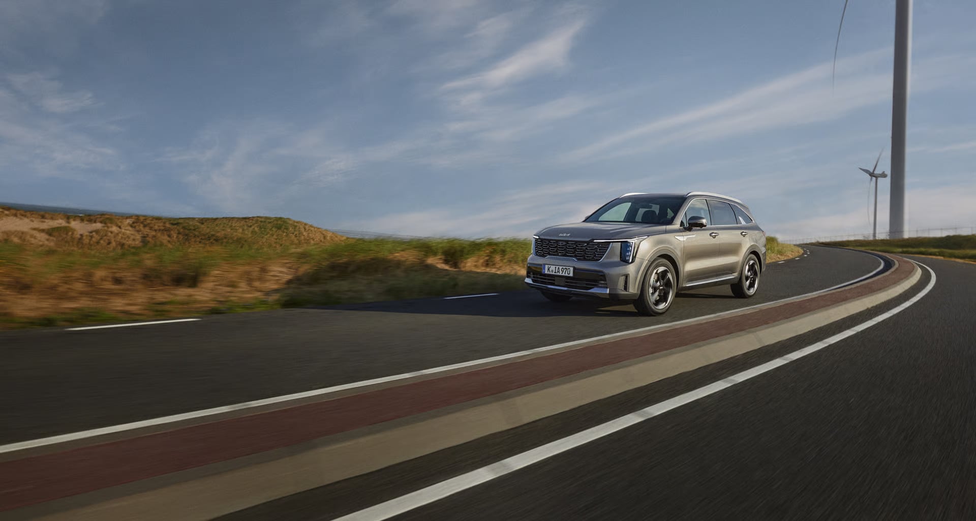 Auf diesem Bild ist ein grauer SUV zu sehen, der auf einer kurvigen Straße fährt, umgeben von begrünten Dünen und Windrädern im Hintergrund.