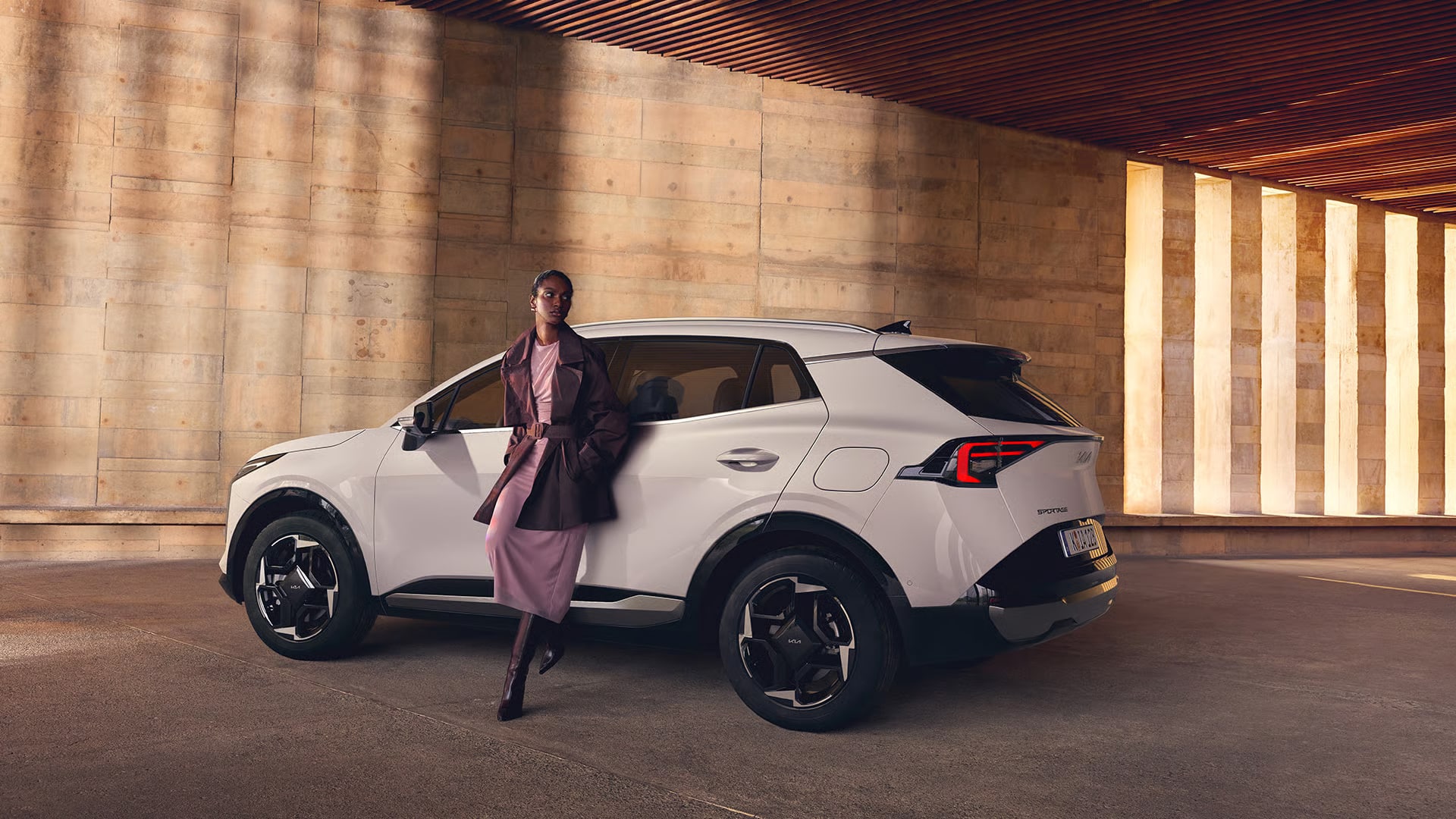 Auf diesem Bild ist eine elegant gekleidete Frau zu sehen, die an einem weißen SUV in einer modernen Architekturkulisse mit warmen Lichtakzenten lehnt.