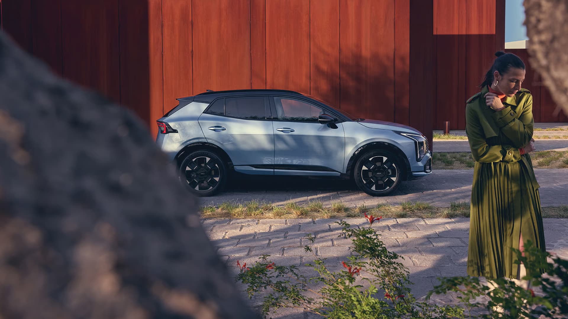 Auf diesem Bild sind ein hellblauer SUV und eine Frau in einem olivgrünen Outfit vor einer großen rostfarbenen Wand, umgeben von Pflanzen im Vordergrund, zu sehen.