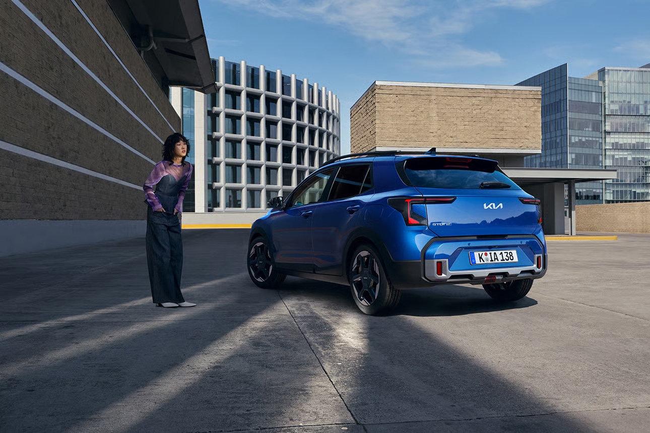 Auf diesem Bild sind ein blauer Kia Stonic und eine danebenstehende Frau vor einer Kulisse aus modernen städtischen Gebäuden zu sehen.