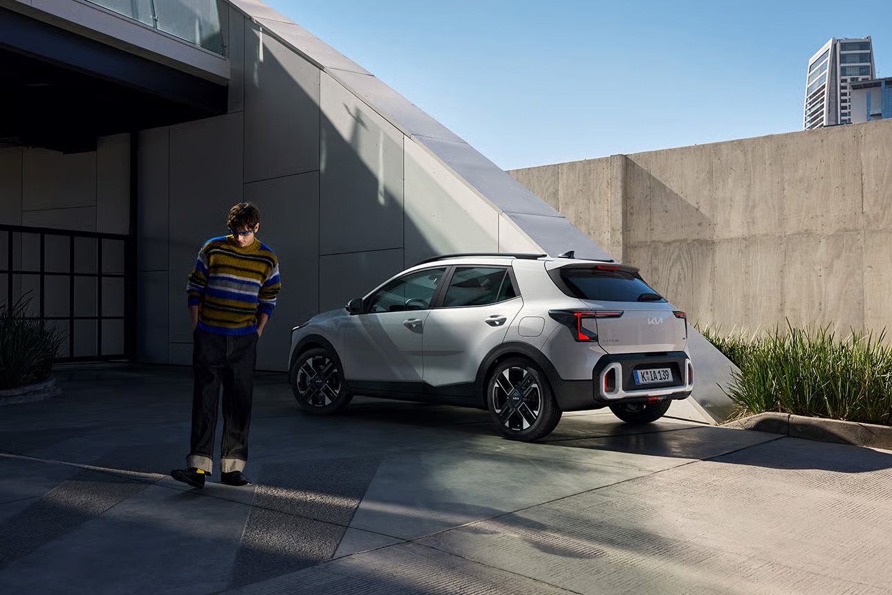 Auf diesem Bild ist ein junger Mann mit einem gestreiften Pullover zu sehen, der neben einem modernen, silbernen Kia in einer urbanen Umgebung mit markanter Architektur steht.