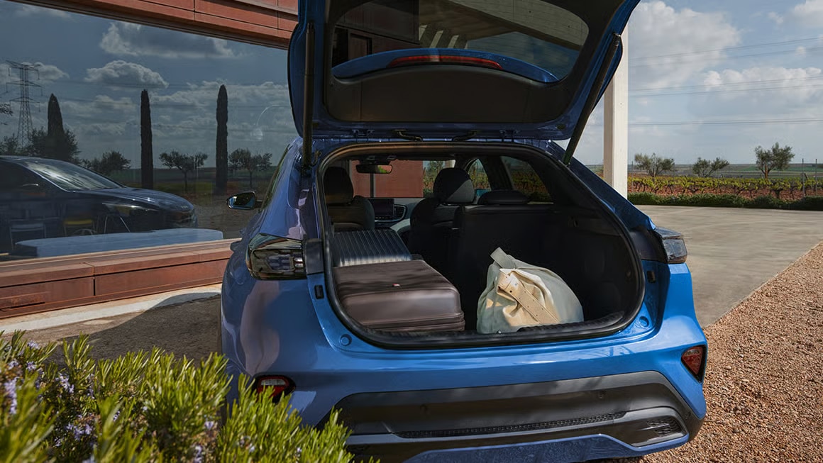 Auf diesem Bild ist ein blaues Auto von hinten mit geöffnetem Kofferraum zu sehen, in dem ein Koffer und eine Tasche liegen, geparkt vor einem modernen Gebäude mit einer Landschaft und einem Weinberg im Hintergrund.