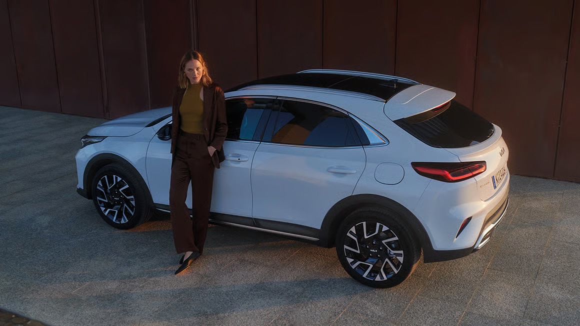 Das Bild zeigt eine Frau in einem braunen Anzug, die an ein weißes Kia SUV gelehnt ist.