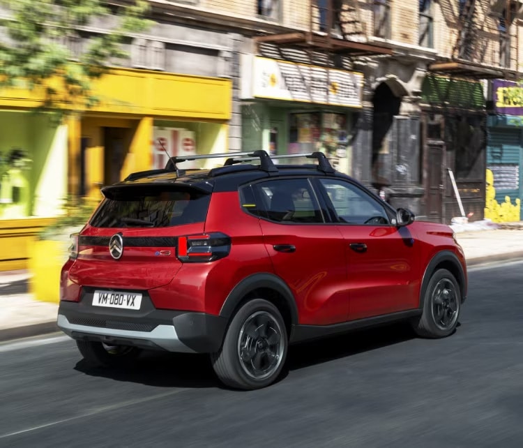 Auf diesem Bild ist ein rotes Auto mit schwarzem Dach zu sehen, das auf einer Straße durch eine städtische Umgebung fährt.