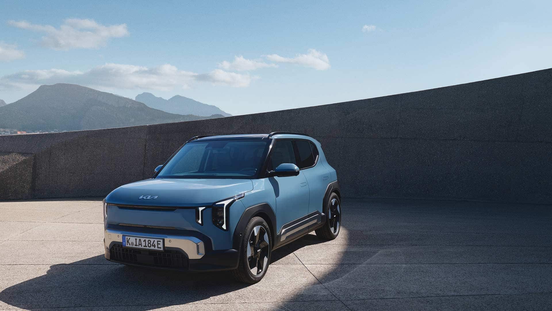 Blauer Kia EV2 Elektro-SUV in der Dreiviertel-Frontansicht auf einem sonnigen Parkplatz vor Bergkulisse.