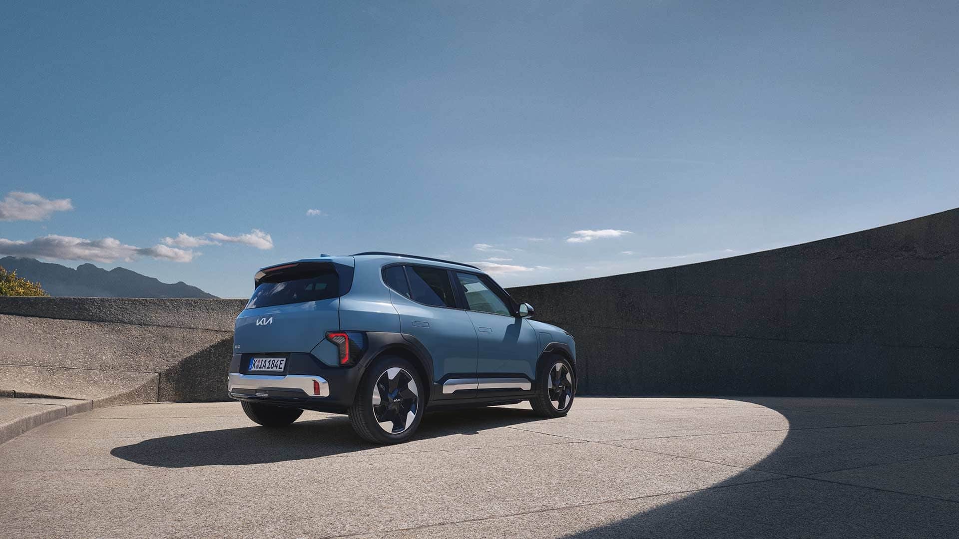 Blauer Kia EV2 Elektro-SUV in der Heckansicht auf einem sonnigen Platz mit Bergpanorama