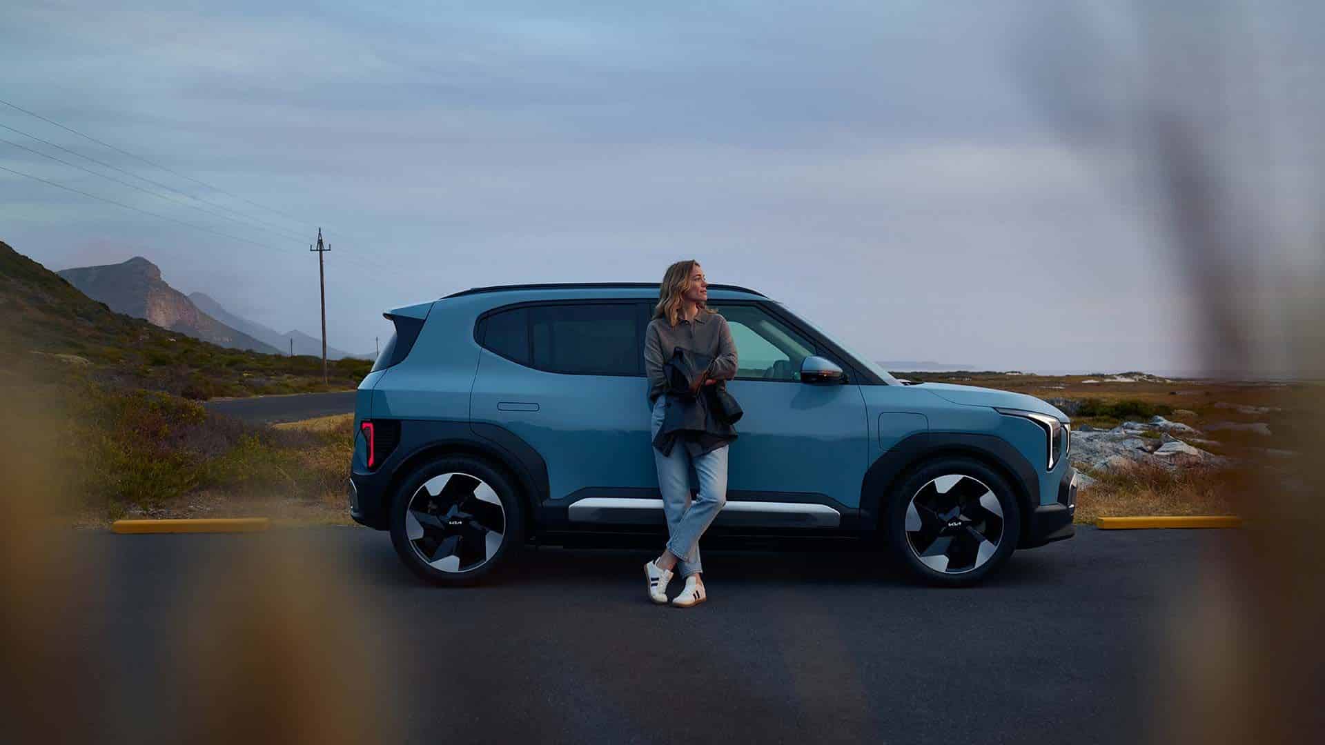 Blauer Kia EV2 Elektro-SUV in der Seitenansicht mit einer Person an einer Küstenstraße.